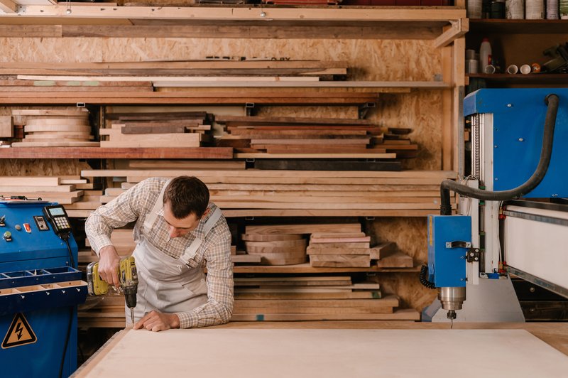 Tradesperson using a power drill on timber in a joinery workshop, with a CNC machine and timber rack in the background