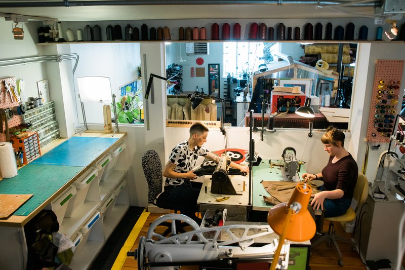 Inside a leather and shoe-repair atelier — two people at sewing machines surrounded by tools and thread
