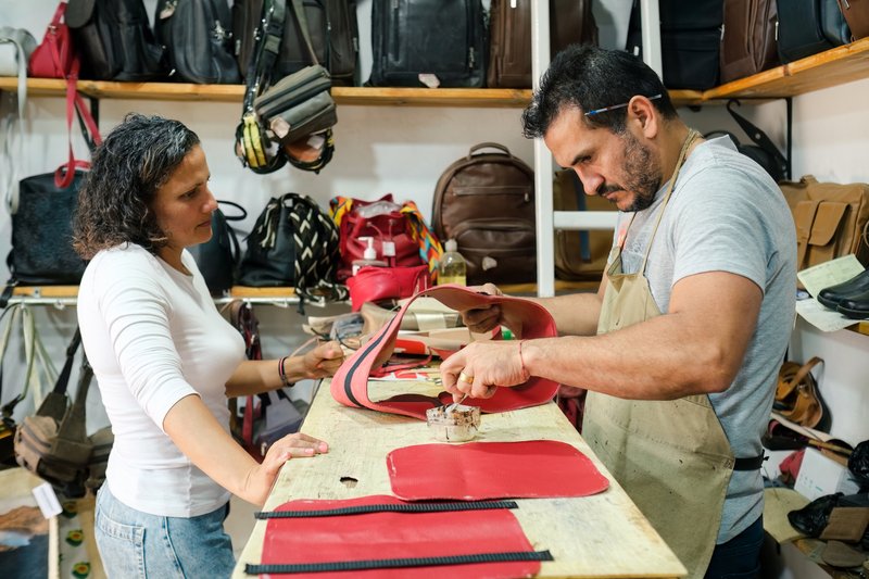 Two leather workers collaborating on a red leather bag in their workshop